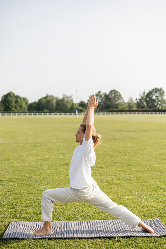 side view of man in linen pants and white t-shirt practicing warrior pose with raised praying hands on yoga mat on green lawn - Photo, Image