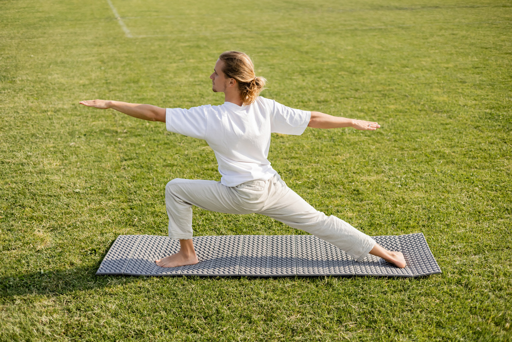 vue latérale du jeune homme aux cheveux longs en t-shirt blanc méditant en posture guerrière avec les mains tendues sur le tapis de yoga à l'extérieur - Photo, image