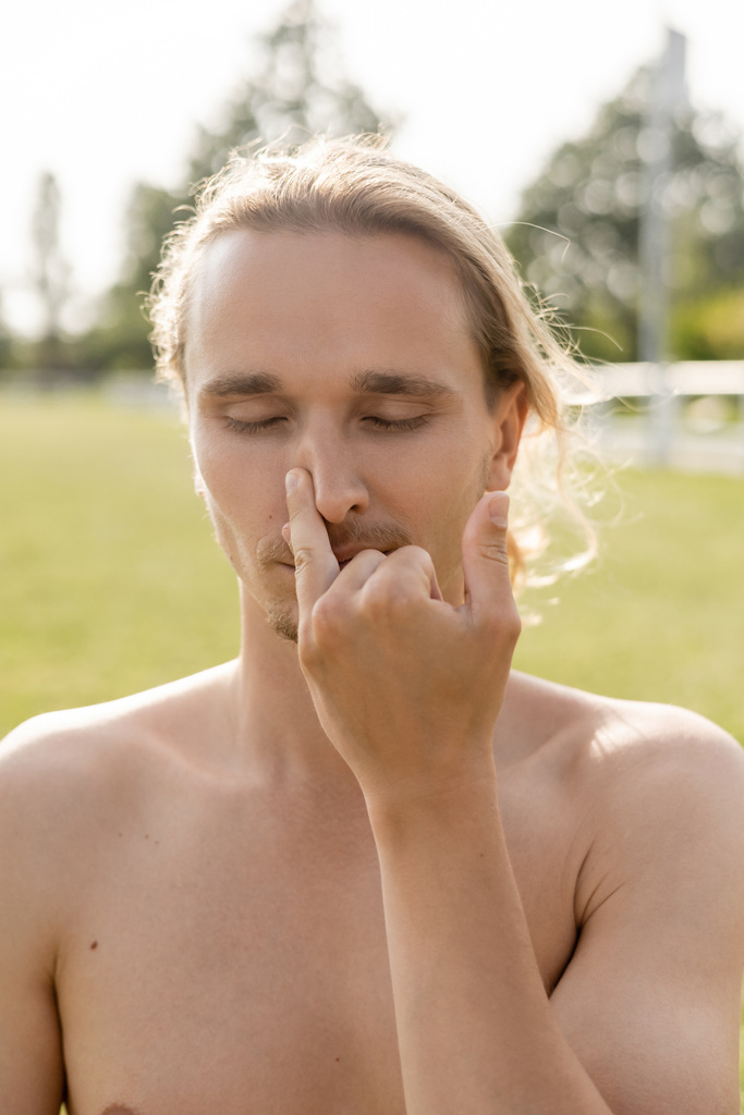 hombre de yoga sin camisa con los ojos cerrados taponando la nariz mientras hace ejercicio pranayama al aire libre - Foto, imagen