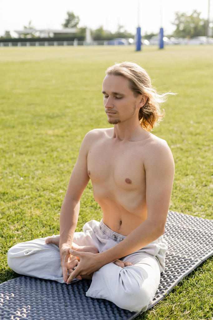 hombre sin camisa de pelo largo en pantalones de lino meditando en pose de loto con los ojos cerrados en el estadio cubierto de hierba - Foto, imagen