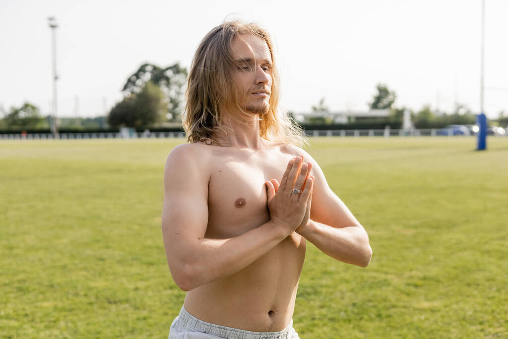 Üstsüz, uzun saçlı yoga adamı çimenli stadyumda dua eden elleriyle meditasyon yapıyor. - Fotoğraf, Görsel
