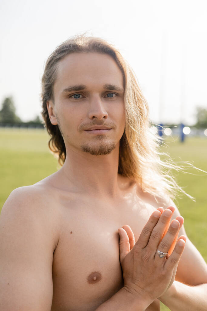 homem sem camisa positivo com cabelos longos meditando com gesto mudra anjali e sorrindo para a câmera ao ar livre - Foto, Imagem