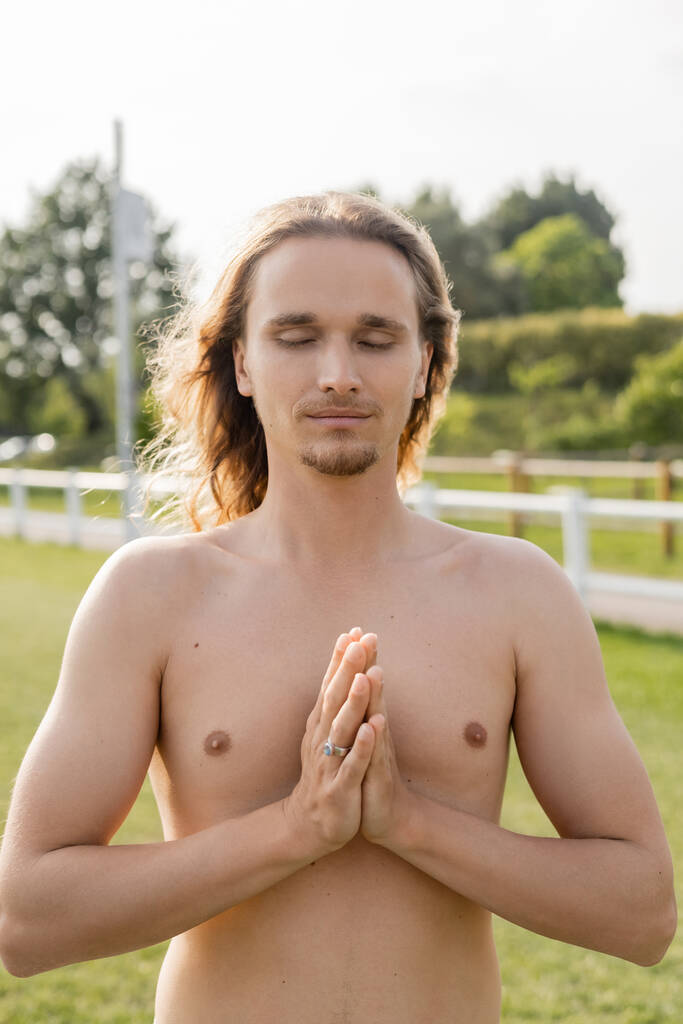 mężczyzna bez koszuli z długimi włosami i zamkniętymi oczami pokazując anjali mudra gest podczas medytacji na świeżym powietrzu - Zdjęcie, obraz