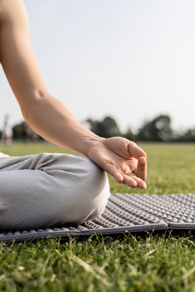 cropped view of young man meditating and showing gyan mudra gesture while sitting on yoga mat on green lawn - Photo, Image