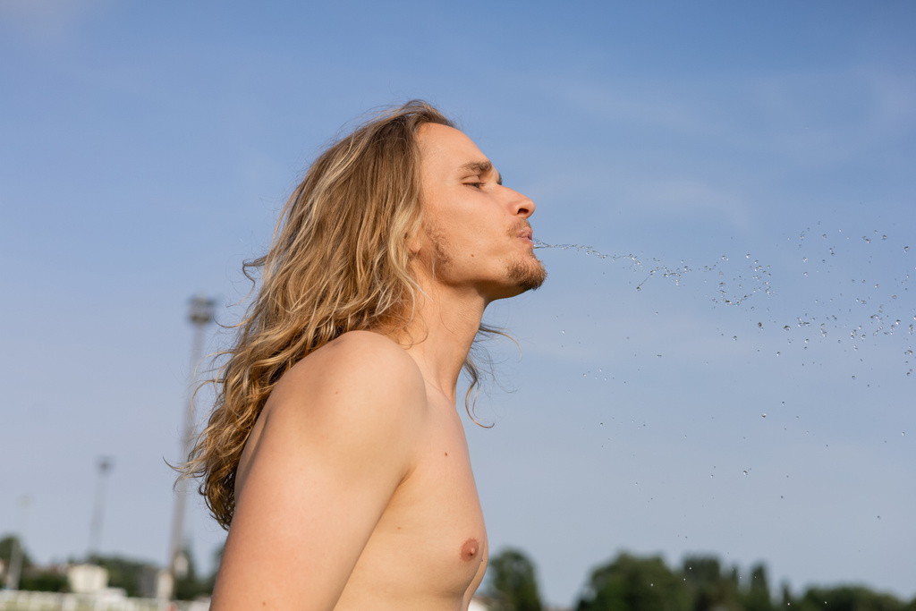 vista lateral do homem de cabelos longos sem camisa cuspir água doce enquanto faz exercício pranayama ao ar livre - Foto, Imagem