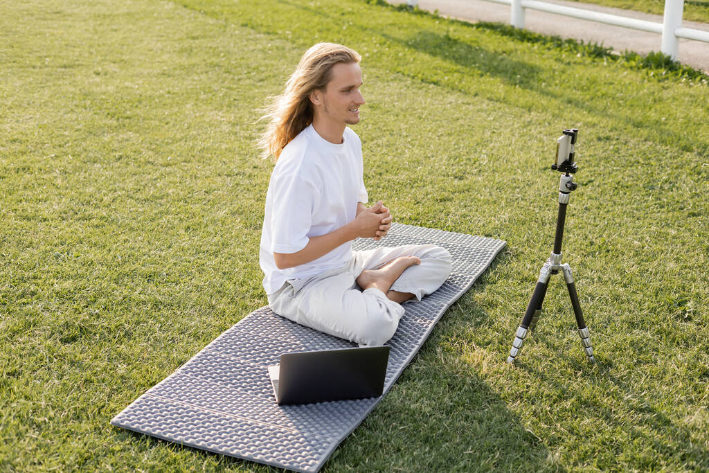profesor de yoga sonriente sentado en pose fácil cerca del trípode con teléfono inteligente y portátil en el césped verde - Foto, imagen