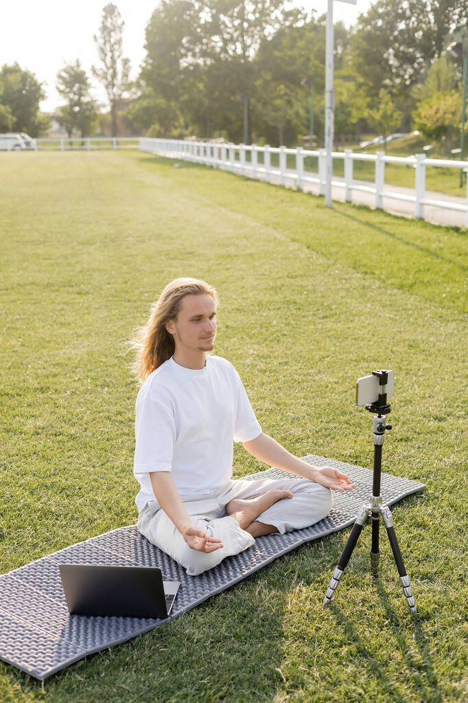 insegnante di yoga mostrando posa facile e gesti mudra gyan mentre seduto su tappetino yoga vicino smartphone su treppiede all'aperto - Foto, immagini