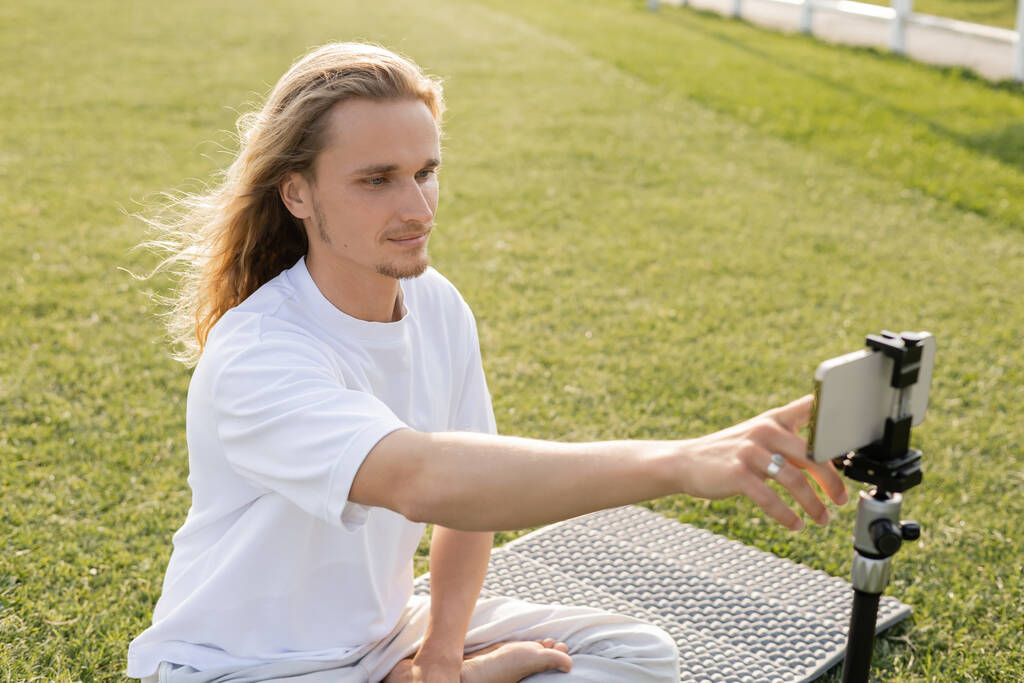 junger langhaariger Mann benutzt Handy, während er auf Yogamatte auf grünem Rasen sitzt  - Foto, Bild
