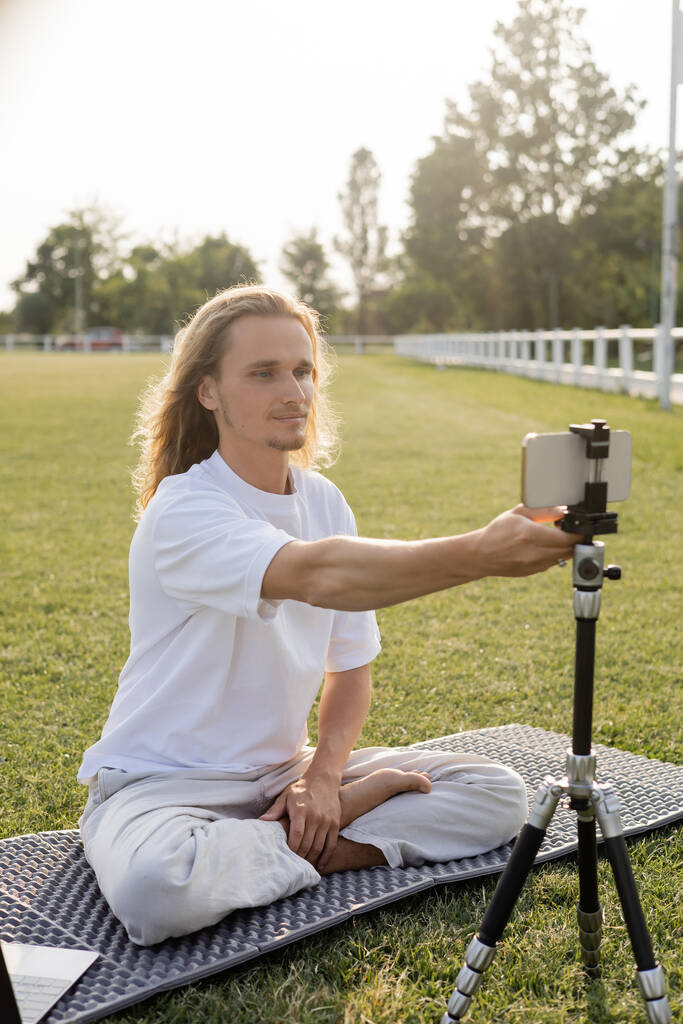 yoga vlogger sitting in easy pose and adjusting tripod with smartphone while sitting on green grass outdoors - Photo, Image