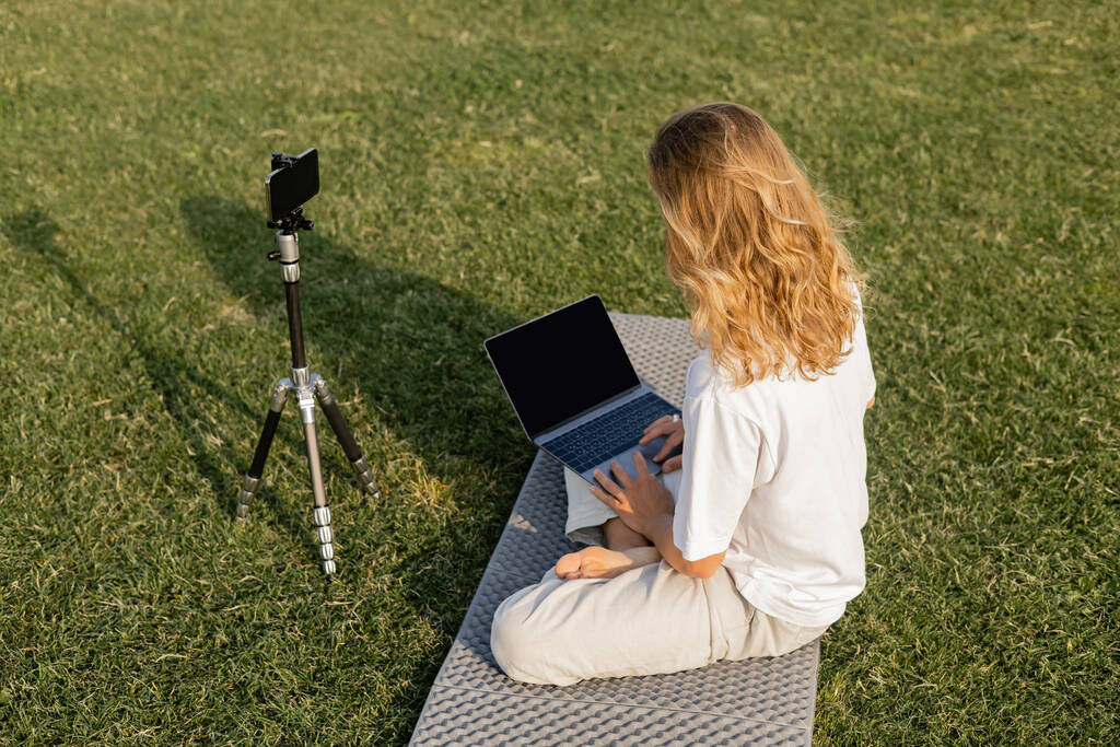 wysoki kąt widzenia długowłosy mężczyzna za pomocą laptopa w pobliżu statywu z telefonu komórkowego podczas lekcji jogi na zielonym polu - Zdjęcie, obraz