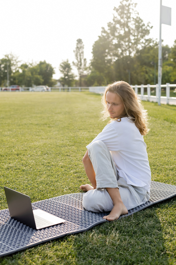 長い髪の男は屋外競技場の緑の芝生のラップトップでオンラインレッスン中にセージポーズに座っている - 写真・画像