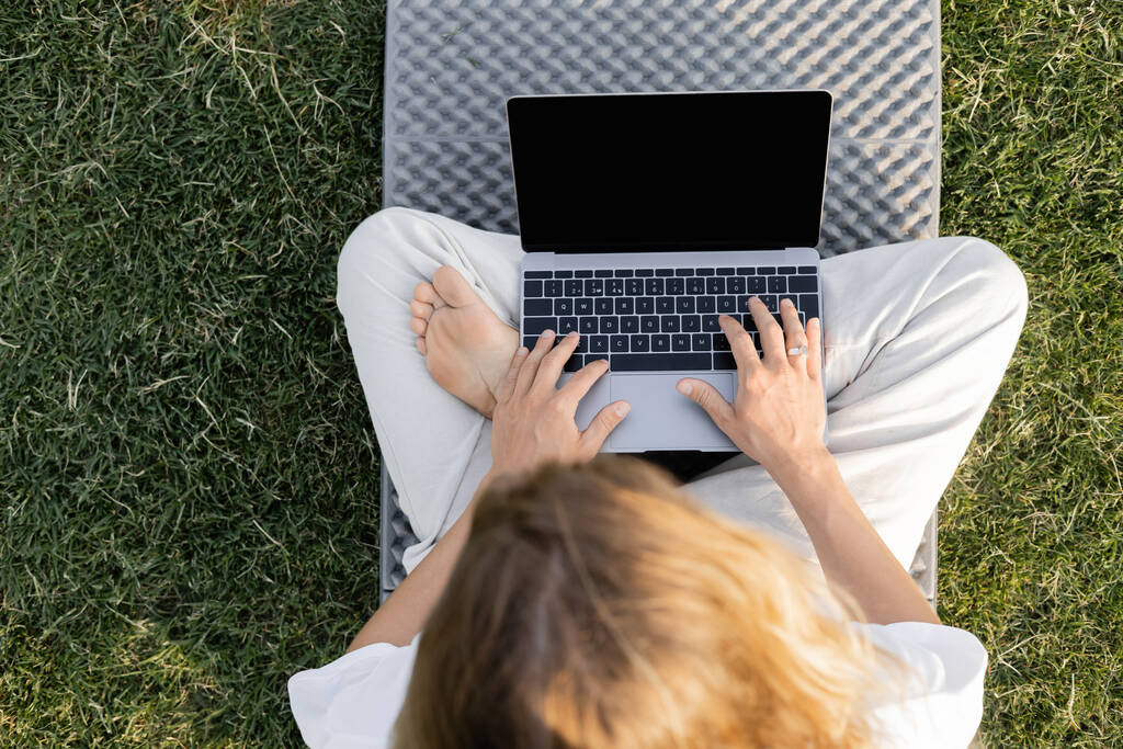 Draufsicht des barfüßigen Mannes, der in lockerer Pose sitzt und Laptop auf Yogamatte auf grünem Rasen benutzt - Foto, Bild
