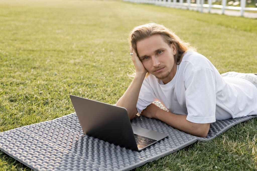 zasněný jóga muž dívá na kameru, zatímco leží v blízkosti notebooku na zelené trávě venku - Fotografie, Obrázek