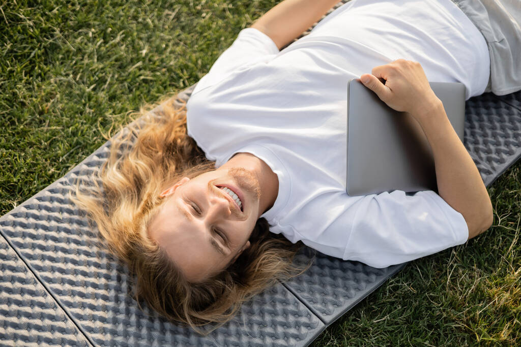 緑の芝生の上でヨガマットの上にノートパソコンを寝そべっている屈託のない長い髪の男のトップビュー - 写真・画像