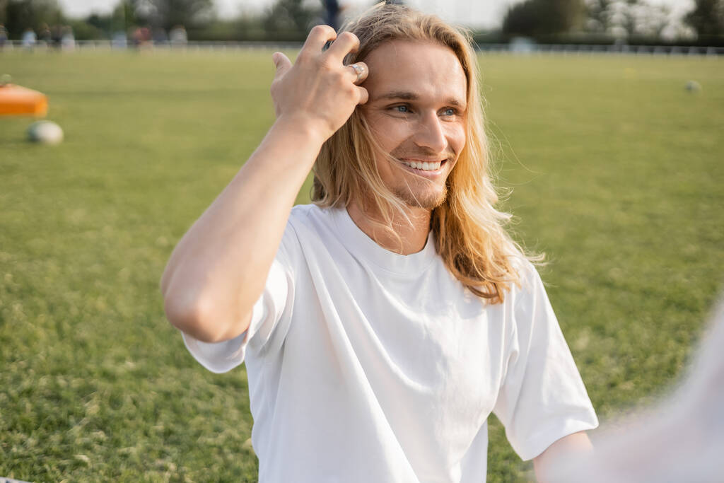 young and carefree yoga man touching long hair and looking away outdoors on blurred foreground - Photo, Image