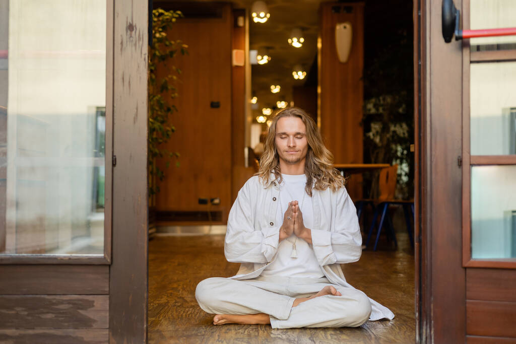 junger langhaariger Mann meditiert in lockerer Pose mit anjali mudra Geste am Hauseingang - Foto, Bild
