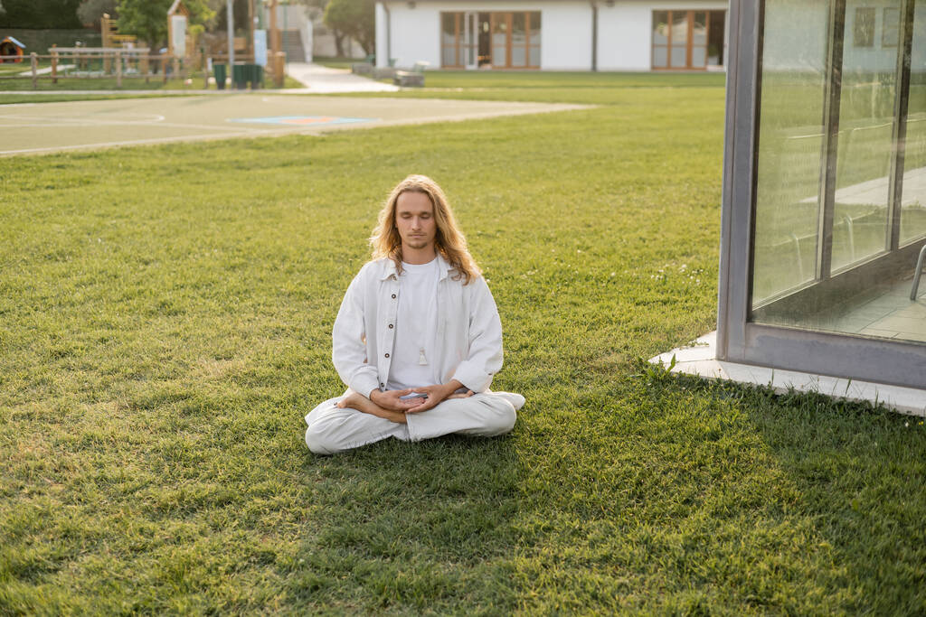 junger und stilvoller Mann in Leinenkleidung sitzt auf dem Rasen in der Nähe von Hütten und meditiert in Lotus-Pose - Foto, Bild