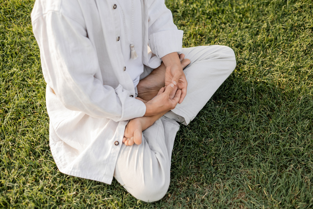 частичный вид босоногого человека в белой льняной одежде медитирующего в позе лотоса на зеленом газоне - Фото, изображение
