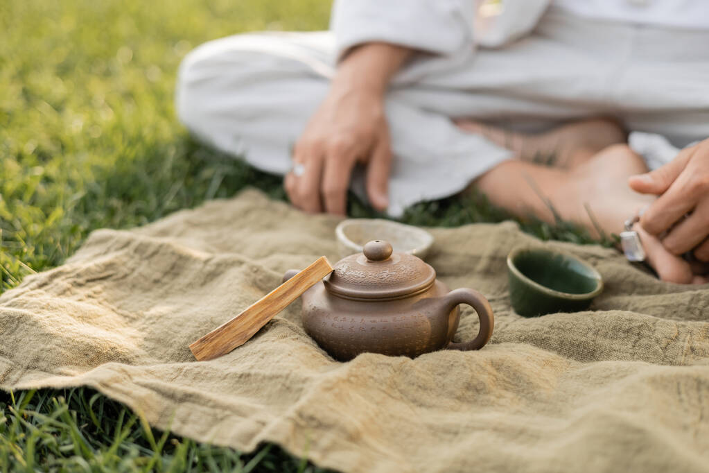 vista parziale dell'uomo yoga seduto vicino al tappeto di lino con palo santo bastone e teiera di argilla con tazze sul prato verde - Foto, immagini