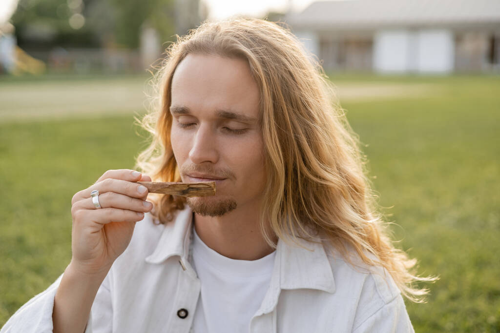 homem de ioga de cabelos longos com olhos fechados desfrutando de sabor de palo santo vara ao ar livre - Foto, Imagem