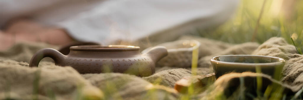 λινό χαλί με πήλινη τσαγιέρα και κύπελλα κοντά περικοπεί άνθρωπος γιόγκα σε θολή φόντο, πανό - Φωτογραφία, εικόνα
