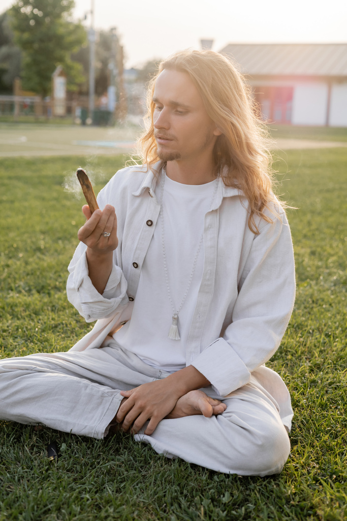 stylish man in white linen clothes sitting in easy pose and holding smoldering palo santo stick while meditating outdoors - Photo, Image