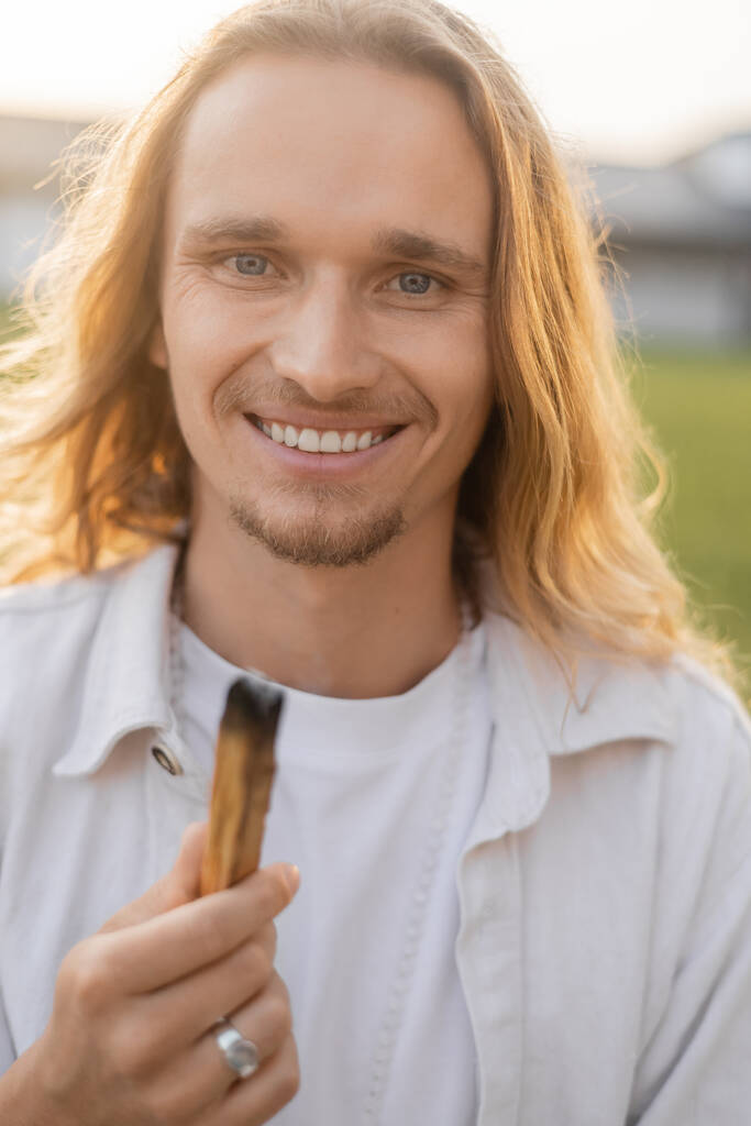 joyful long haired yoga man holding smoldering palo santo stick and smiling at camera outdoors - Photo, Image