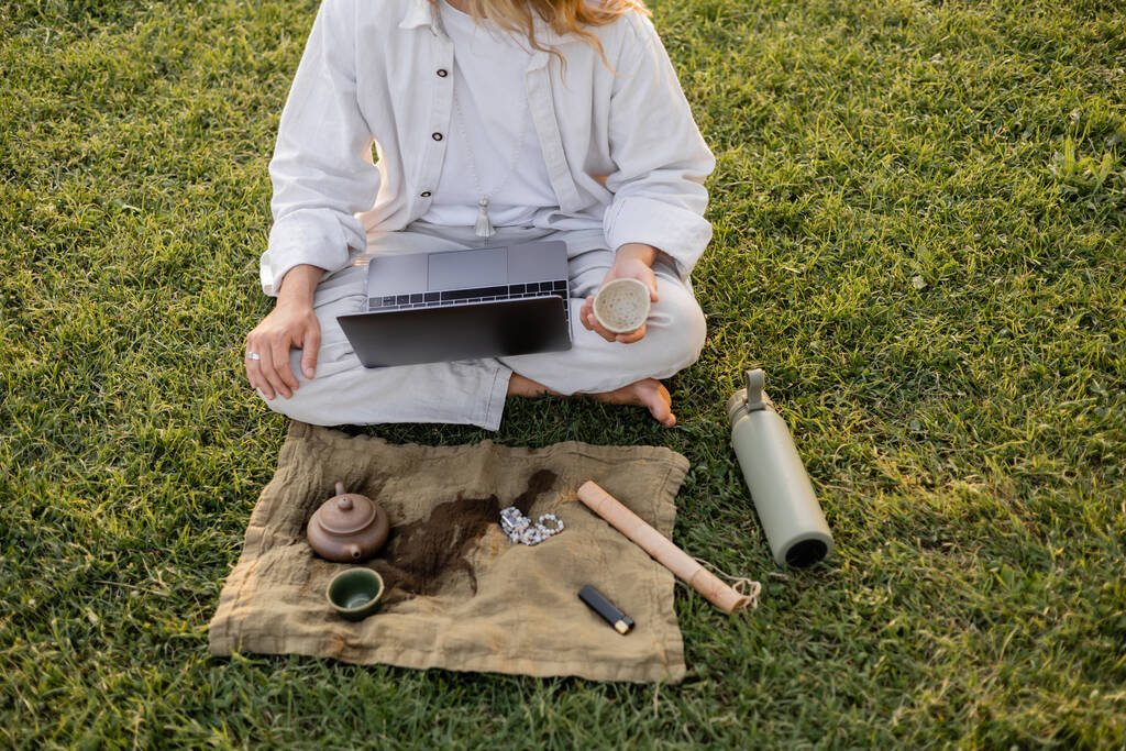 gedeeltelijk zicht op yoga man zittend op groen gazon met laptop en puer thee in de buurt van thermos en linnen tapijt met klei theepot en mala kralen - Foto, afbeelding