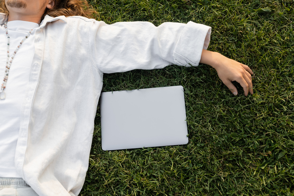bovenaanzicht van bijgesneden man in wit katoenen shirt en kralen liggend op groen gras in de buurt van laptop - Foto, afbeelding