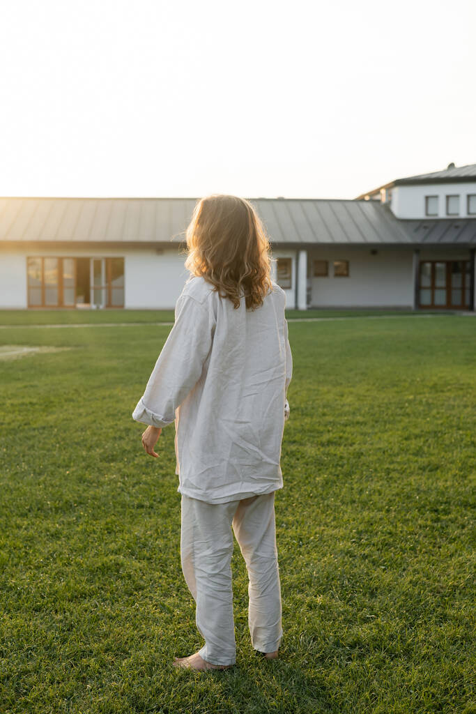 back view of barefoot and long haired man in linen shirt and pants standing on green lawn near countryside house - Photo, Image