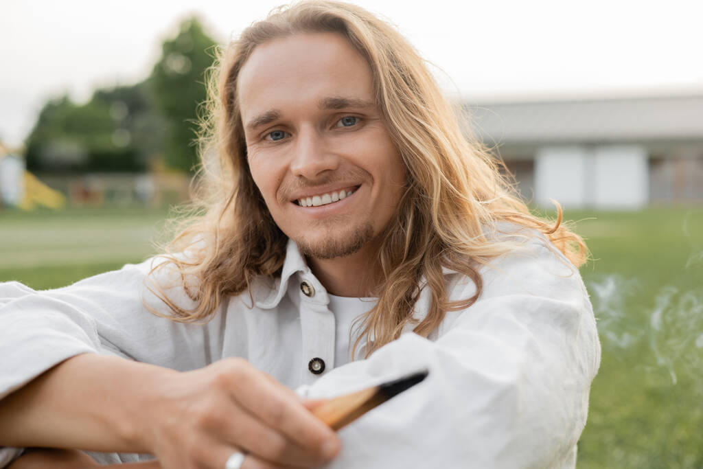 carefree long haired man holding fragrant palo santo stick and looking at camera outdoors - Photo, Image