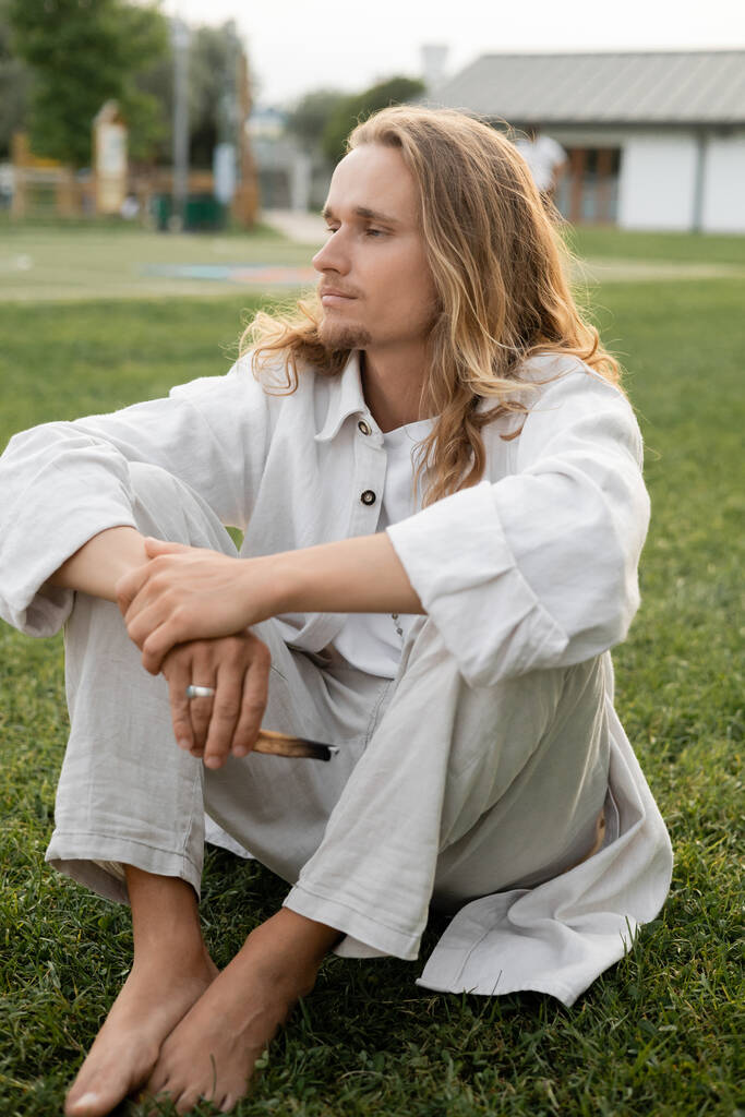 ruhiger, barfüßiger Mann in weißen Leinenkleidern, der beim Sitzen mit aromatischem Palo Santo im Freien wegschaut - Foto, Bild