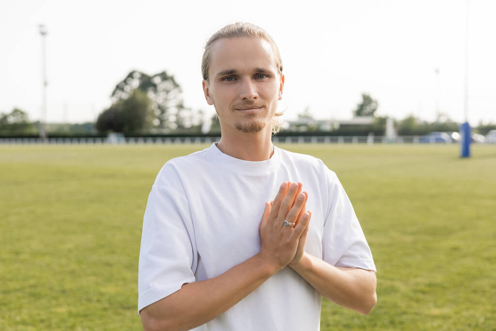 hombre joven y positivo en camiseta blanca que muestra el gesto de anjali mudra y mirando a la cámara en el estadio al aire libre borrosa - Foto, imagen