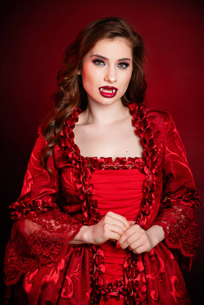 Retrato de uma jovem e atraente mulher vampira em um vestido rococó vermelho posando isolada contra um fundo escuro com backlights vermelhos. - Foto, Imagem