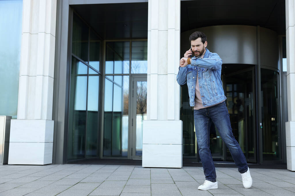Akıllı telefondan konuşan mutsuz bir adam binanın yanındaki saate bakıyor. Geç kalma kavramı - Fotoğraf, Görsel