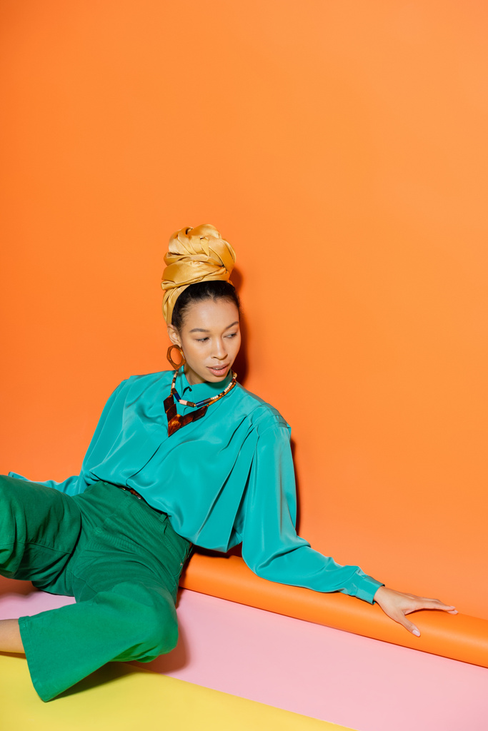 Türbanlı ve mavi bluzlu, turuncu arka planda oturan modaya uygun Afrikalı Amerikalı kadın.  - Fotoğraf, Görsel