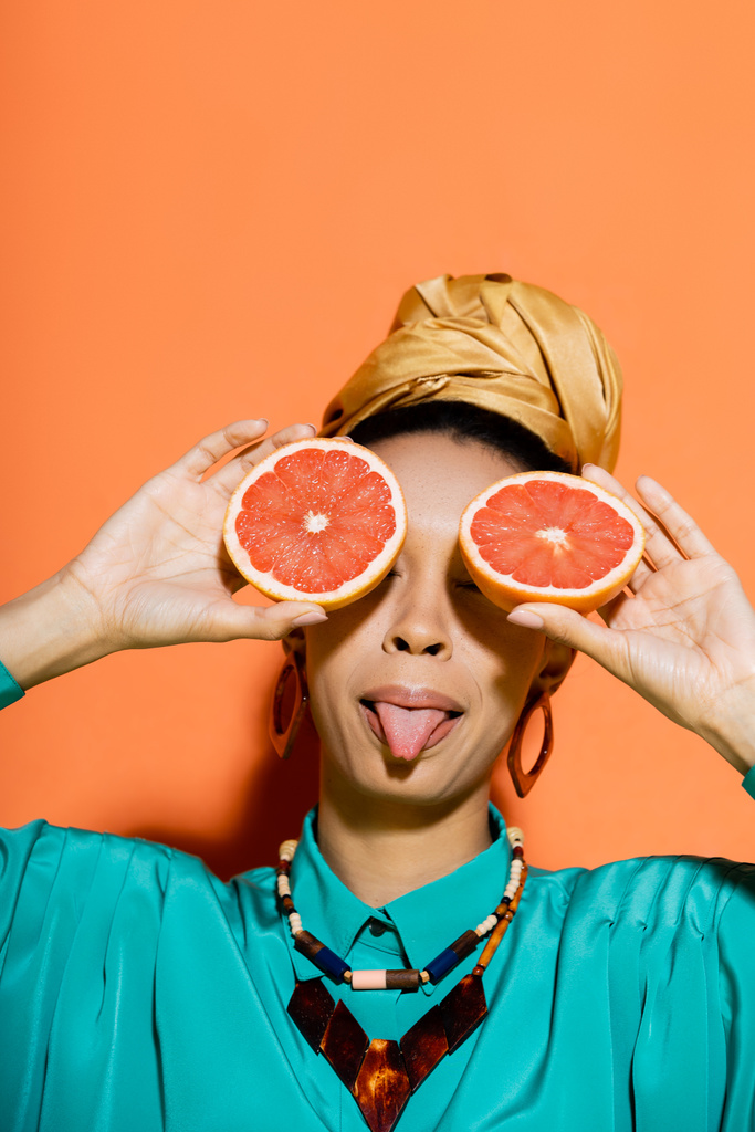 Elegante mujer afroamericana sobresaliendo lengua y sosteniendo toronja cortada sobre fondo naranja  - Foto, Imagen