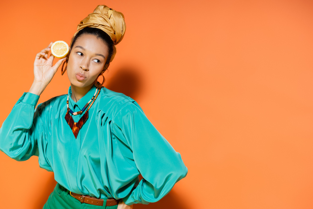Κομψό αφροαμερικάνικο μοντέλο που μουτρώνει τα χείλη και κρατάει κομμένο λεμόνι σε πορτοκαλί φόντο  - Φωτογραφία, εικόνα