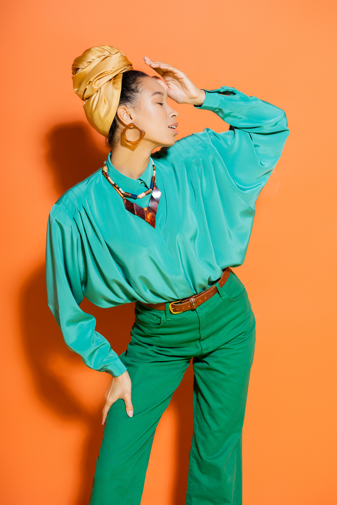 Vista lateral de la modelo afroamericana de moda posando en ropa de verano sobre fondo naranja  - Foto, Imagen