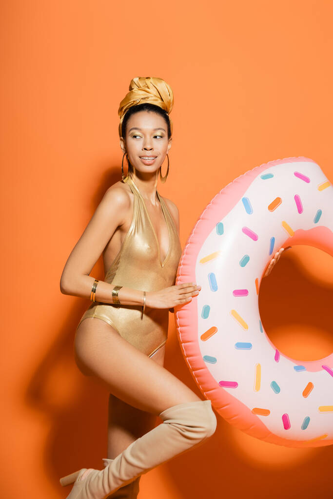 Улыбающаяся африканская американка в золотом купальнике держит кольцо в бассейне на оранжевом фоне  - Фото, изображение