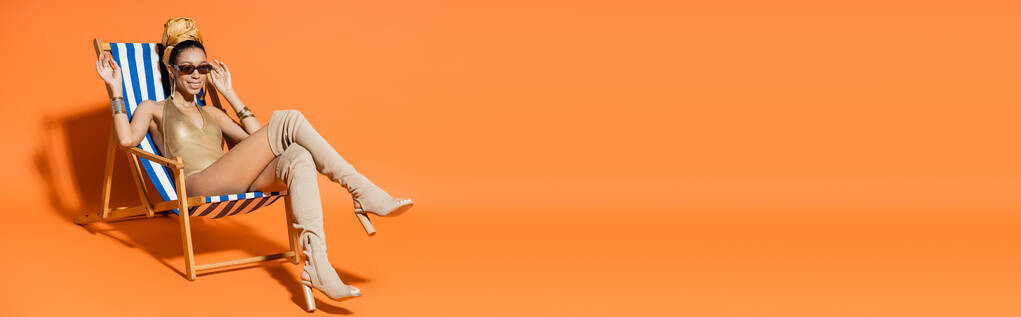 Trendy e sorridente donna afroamericana in costume da bagno sventolando mano mentre seduto sulla sedia a sdraio su sfondo arancione, banner  - Foto, immagini