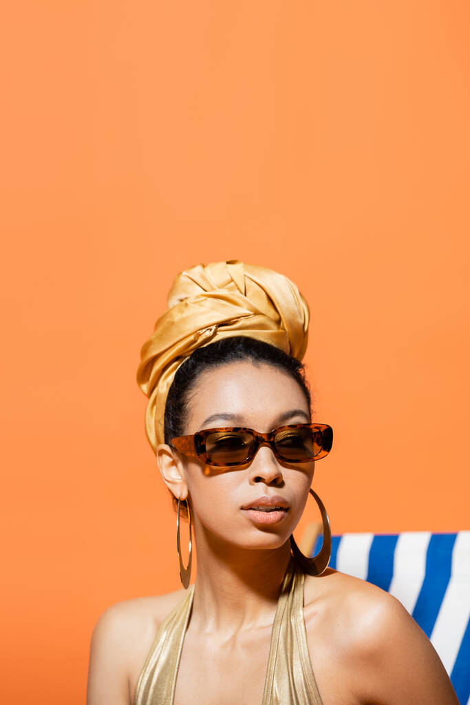 Türbanlı ve güneş gözlüklü modaya uygun Afro-Amerikan modelinin portresi portakallı güverte sandalyesinin yakınına bakıyor.  - Fotoğraf, Görsel