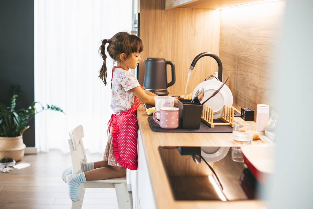 Cute little girl washing dishes with sponge standing on the chair. Child doing chores concept. - Photo, Image