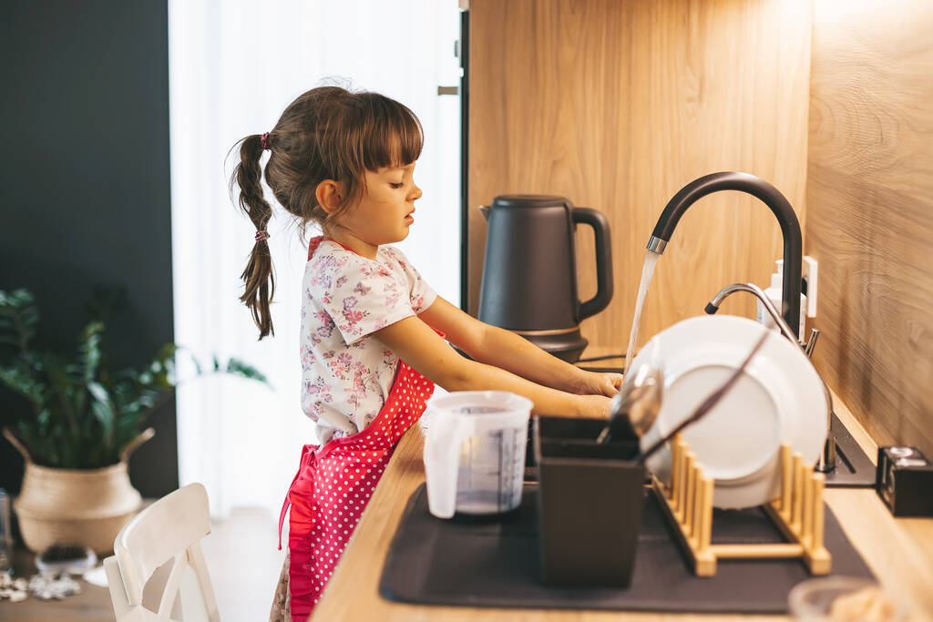 Mała dziewczynka zmywa naczynia gąbką w zlewie kuchennym w domu. Koncepcja wykonywania obowiązków przez dziecko. - Zdjęcie, obraz