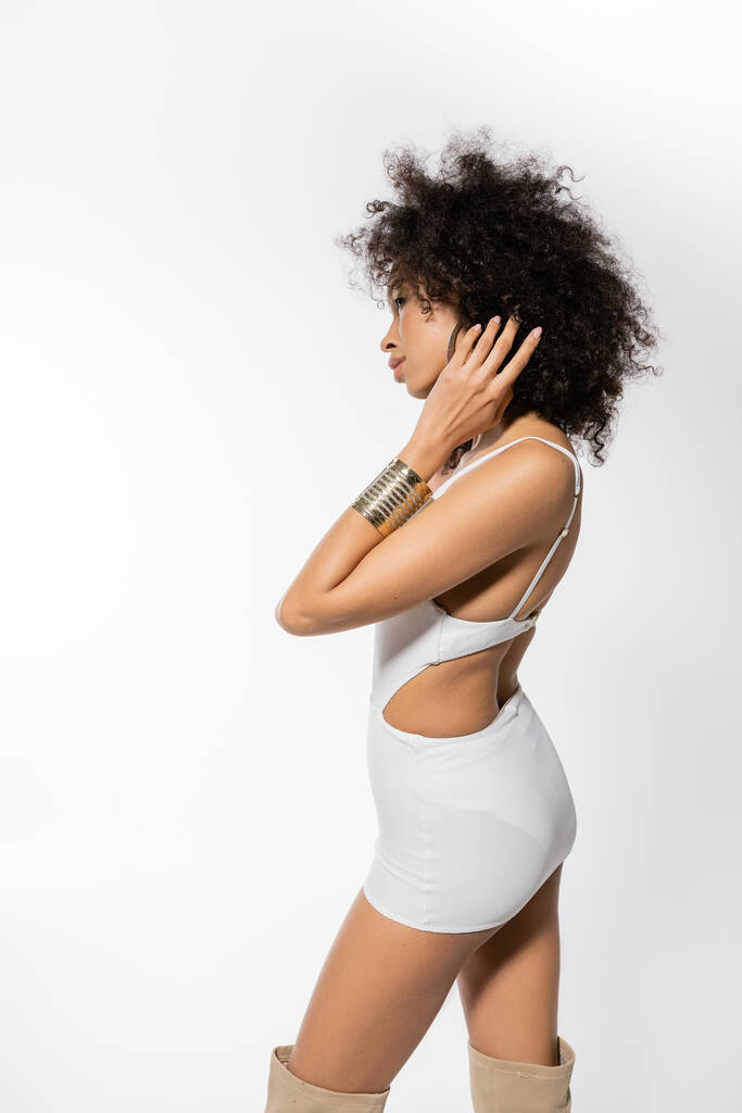 widok z boku młody afrykański amerykański model z kręcone włosy pozowanie w biały mini sukienka na szarym tle - Zdjęcie, obraz