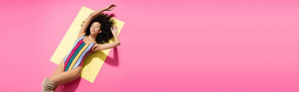 вид сверху на кудрявую африканскую американскую модель в красочных полосатых купальниках, лежащих на желтом и розовом фоне, баннер  - Фото, изображение