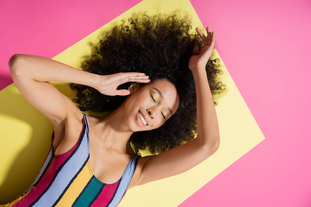 top uitzicht van blij Afrikaans Amerikaans model in kleurrijke gestreepte zwembroek liggend op gele en roze achtergrond  - Foto, afbeelding