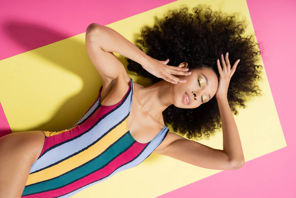 vrchní pohled na bezstarostný africký americký model v barevných pruhovaných plavkách ležících na žlutém a růžovém pozadí  - Fotografie, Obrázek