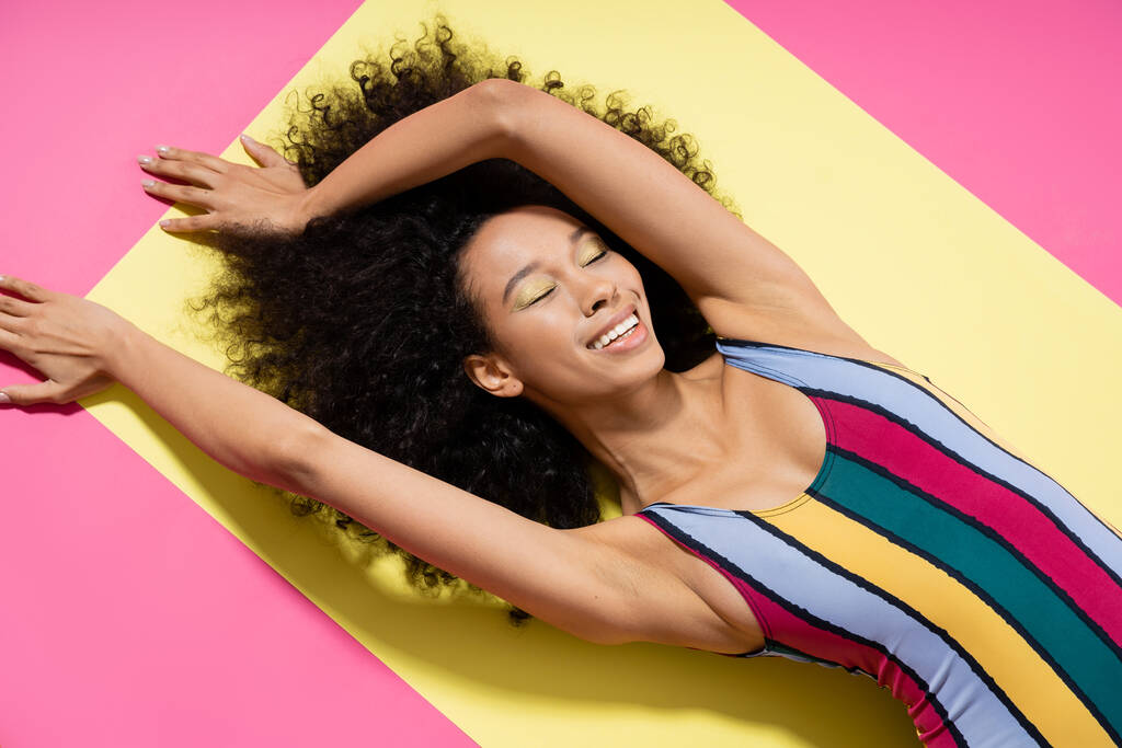 黄色とピンクの背景に日焼けを取得カラフルな水着で幸せなアフリカ系アメリカ人女性のトップビュー  - 写真・画像