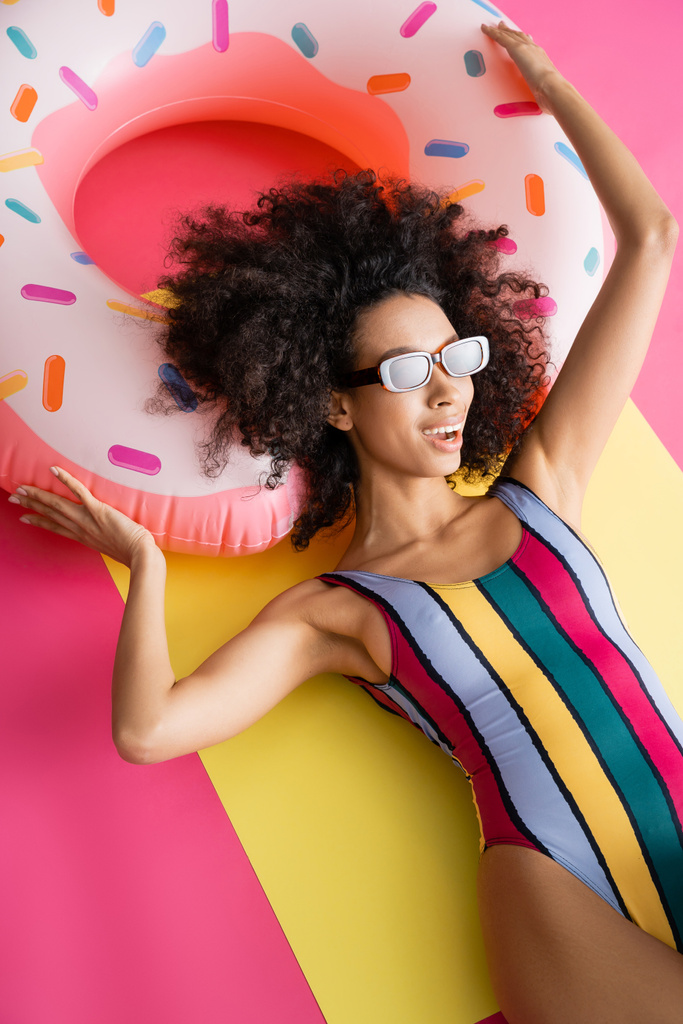 κορυφαία άποψη του θετικού αφρικανικού αμερικανικού μοντέλου σε ριγέ μαγιό και γυαλιά ηλίου που μαυρίζουν κοντά σε φουσκωτό δαχτυλίδι σε κίτρινο και ροζ  - Φωτογραφία, εικόνα